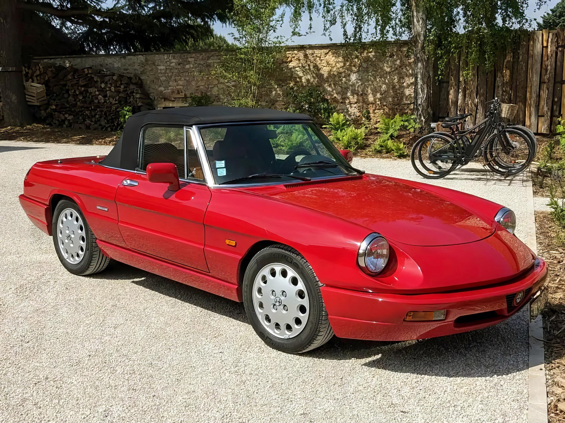 La voiture Alpha Romeo Spider rouge décapotable, qu'il est possible de louer lors de votre séjour à la maison d'hôtes Val Trebbia, garée sur une allée gravillonnée, avec des vélos en arrière-plan et un décor naturel entouré d'arbres et d'une clôture en bois.